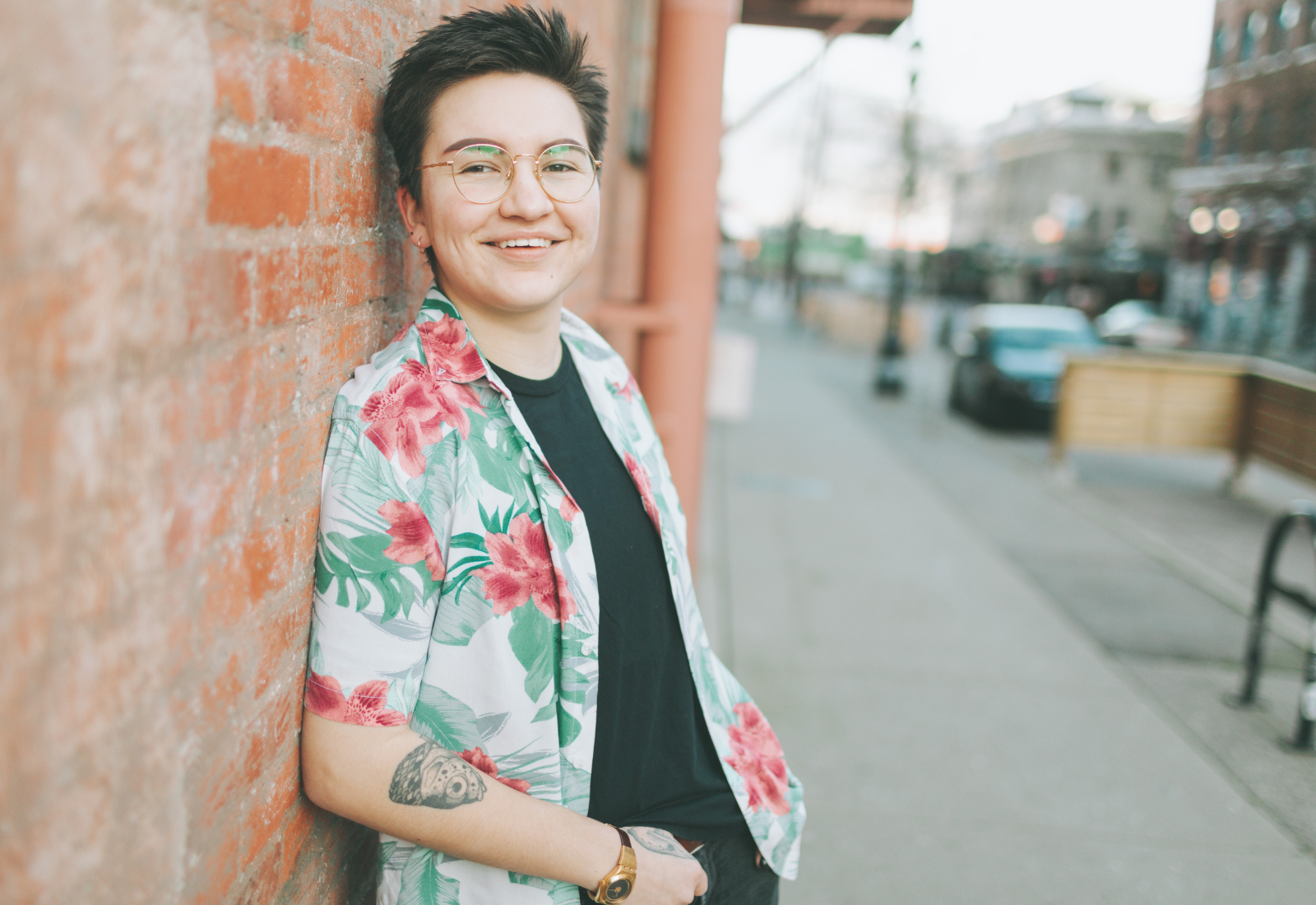 smiling young trans man wearing glasses standing in front of a brick wall